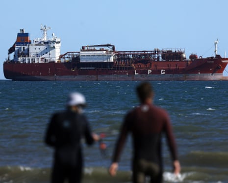 Two people in wetsuits stand on a beach with a large red oil tanker visible offshore