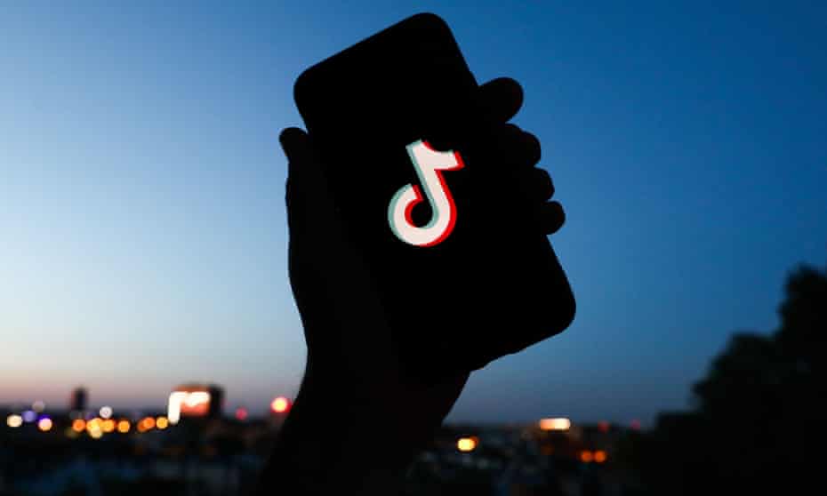 The silhouette of a hand holding a smartphone with the TikTok logo on it is shown against the backdrop of a city skyline at dusk.