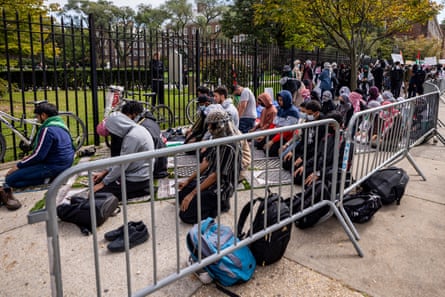 A group of students sit cross-legged on sidewalk between an iron fence (next to a green lawn) and a police fence.