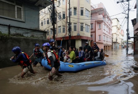 Three men wearing helmets pull a small blue boat full of people through a flooded street