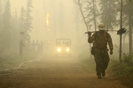 A firefighter, head down and carrying tools, walks down a road through a smoke-filled forest. A vehicle and other firefighters can be seen behind the first.