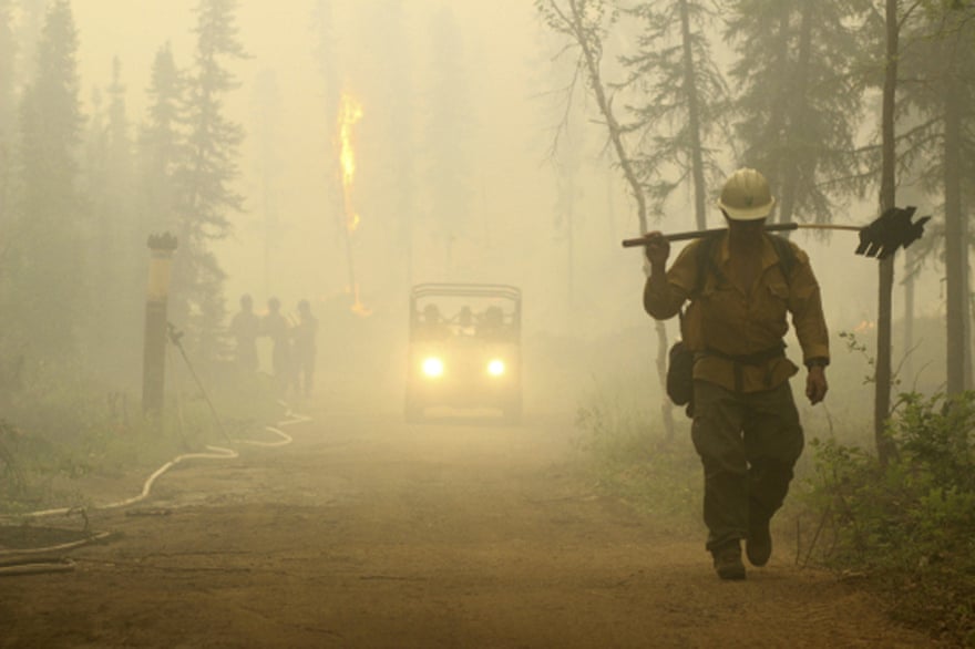 A firefighter, head down and carrying tools, walks down a road through a smoke-filled forest.  A vehicle and other firefighters can be seen behind the first.