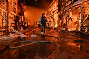 Fire engulfs a market as a firefighter in protective gear walks over hoses as colleagues in the background tackle the blaze