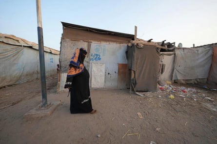 A fully covered woman stands on dusty ground outside a small makeshift shanty