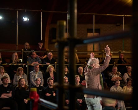 Ian McKellen holds his hands above his head as he speaks. An audience looks on behind him.