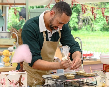 Aaron, in a green jumper and brown apron, pipes icing on to biscuits on a cooling tray.