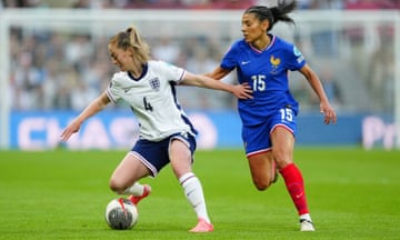 Two female footballers on a green pitch: England's Keira Walsh, in a white shirt and dark blue shorts, who has the ball at her feet, tries to evade France's Kenza Dali, behind her in royal blue with red socks.