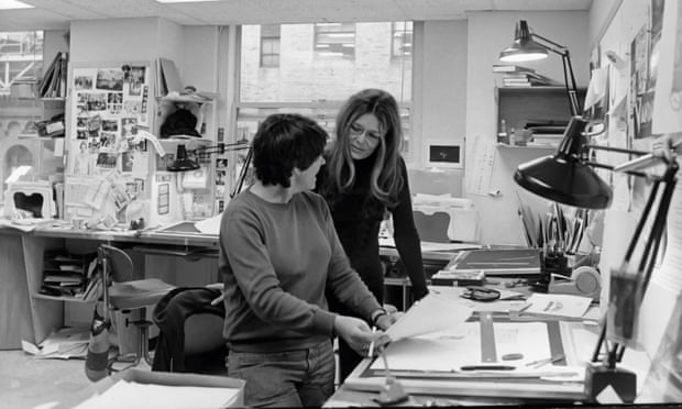 Steinem and colleague look at pages over cluttered desk with anglepoise lamps. Photographs pinned on wall behind