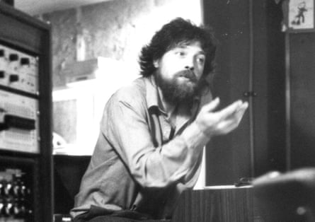 Fay, in a shirt and with a beard, holds out his hand next to recording equipment in a studio in the 1970s