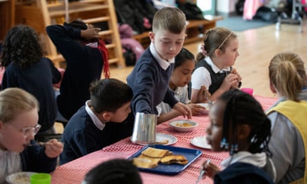 Children at a breakfast club at a primary school in the West Midlands: a group of boys and girls from different ethnic backgrounds sit at a table covered with a red cloth and take pieces of toast from a large plate.