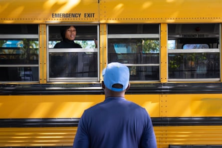 A woman speaks from an open window on a yellow bus to a person standing with their back to the camera