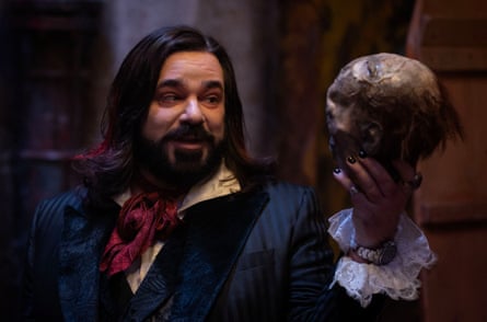 A head and shoulders of a thick-set man with long black hair and a black beard, wearing an Edwardian suit and red cravat, holding a skull aloft as in Hamlet soliloqy - with smile on his face