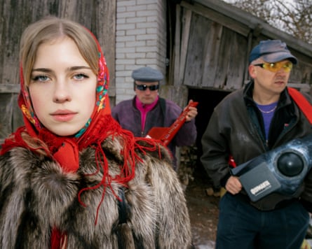 A woman in a red headscarf and fur coat looks moodily at the camera while two men holding instruments stand behind her.