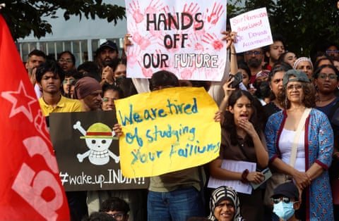 Someone in a crowd holds up a sign that says: ‘We are tired of studying your failures’