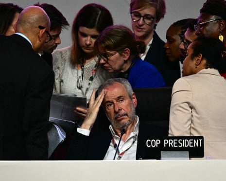 A tired looking man in a suit sits in a chair next to a sign saying 'Cop president', with people gathered in discussion behind him