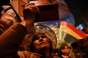 A woman in a wide-brimmed hat takes a photo with her mobile as someone waves a Bolivian flag behind her