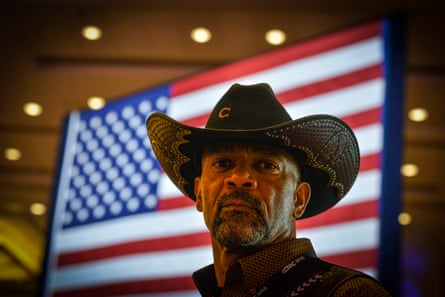 A man in a cowboy hat stands in front of an American flag.