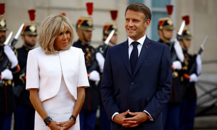 Emmanuel Macron and his wife, Brigitte wearing a white suit, stand next to each other with French guards in the background.