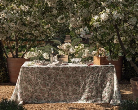 A table set with a floral tablecloth, crockery and napkins, with flowering trees behind it