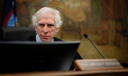 Older white man with poofy white hair wearing black judge’s robe sits at big wooden desk with laptop.