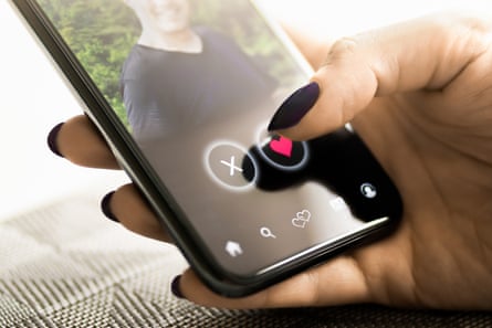 A woman’s thumb clicks the icon of a heart on her phone which is showing a man’s profile photo