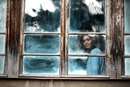 A woman stands side-on, gazing out of a wooden-framed casement window
