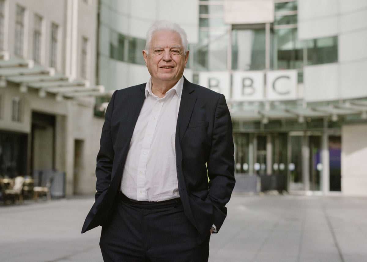 A man with white hair wearing a dark suit and white shirt, standing in front of the BBC building.