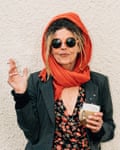 Mid portrait of a woman with blond dreadlocks and a bright orange headscarf and a cigarette in hand and a dark green jackets and orange and black flowery print dress