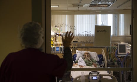 A woman holds up her hand to a window in greeting. On the other side of the window is an elderly man in a hospital bed who has been hooked up to monitoring equipment.