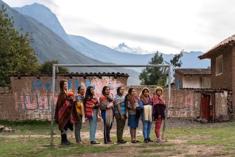 Eight women pose in a goalmouth, wearing traditional woven shawls, with mountain peaks in the background