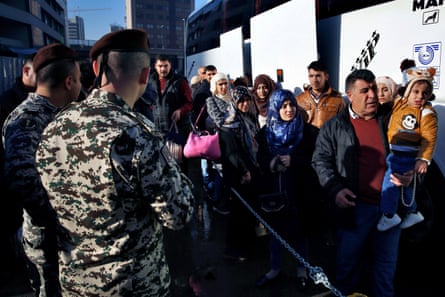 Men in uniform watch a line of people, including children, in a queue to board a bus.