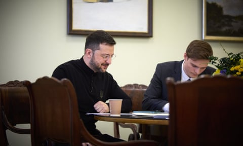 Volodymyr Zelenskyy sitting at at desk with an aid sitting next to him, writing notes