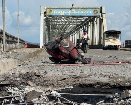 A burnt motorcycle lies on the broken concrete road of a bridge with, behind it, a parked coach and a man in a vest driving a motorbike