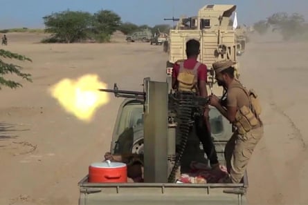 Two fighters fire a machine gun from the back of a pickup truck on a sandy track. Other pickups and armoured vehicles are ahead.