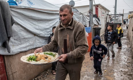 Man, with dish of food, and children walking through mud at Debaga camp