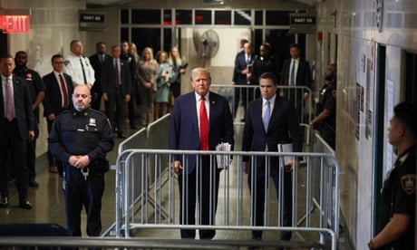 Standing in a sort of rectangular cage of metal police gates, with an armed guard just outside of it, an older white man next to another white man, both in suits, appears to speak. It's all in a dim, narrow corridor.