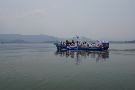 A group of about 20 people raise flags as they sit on a small boat going across a lake with hills in the background.