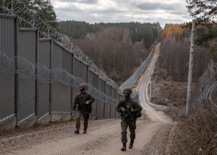 Two armed soldiers patrol on a path beside a tall metal fence with coils of barbed wire on the top; the path stretches downhill behind them through a forest. This is the Polish border with Belarus in the Podlasie region, where fortifications will be aimed at preventing any future Russian invasion.