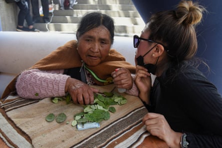 A young woman leans towards an older Indigenous Andean woman who has leaves spread out on a platform before her, partly obscuring some banknotes