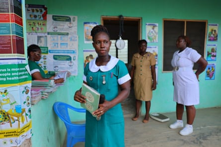 Four nurses in different uniforms stand in a concrete-floored, green-painted clinic covered in health posters.