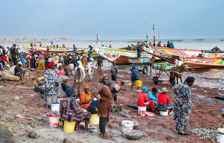 African women in headscarves and colourful print dresses stand on a beach as brightly painted long wooden fishing boats are pulled up