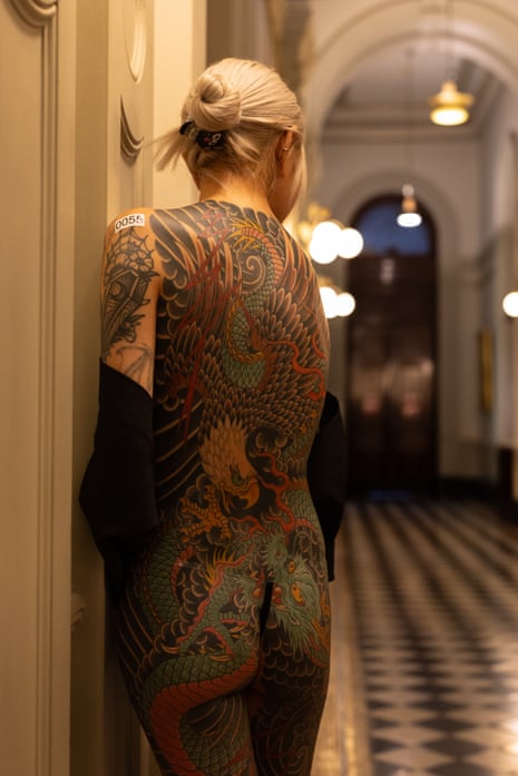 Shell, with blond hair pulled back into a knot stands in a lack and white tiled hall with her back to camera, showing a full body tattoo