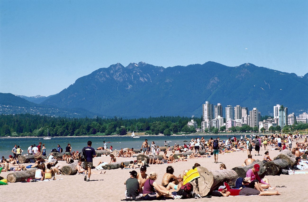 Visitors to a beach at the edge of a big city crowd together in the sand next to carefully placed rows of logs.