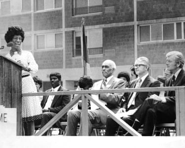 A woman speaks into a microphone as seated men listen.