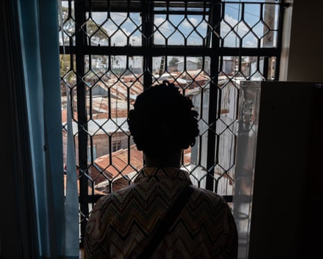 A woman, back to camera, looks out of a window with a metal grille, over the rooftops of the Kibera slum in Nairobi, Kenya.
