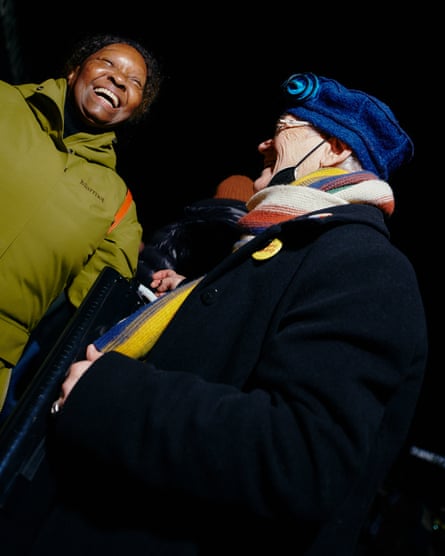 Woman wearing green coat looks forward, laughing, as another woman looks at her, smiling.