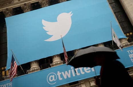 A large sign with the Twitter logo hangs above a banner with the Twitter handle on the front of the New York Stock Exchange building. In the foreground, a person holding a black umbrella is walking by.