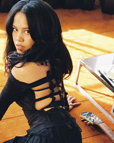 Baby Rose sits on a wooden floor in a black cut-out outfit beside vinyl records and a guitar