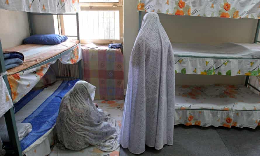 Two women wearing bedsheets as burqas face away from the camera in a room with two three-tier bunk beds