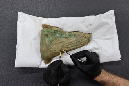 Green bronze object on white cloth with person’s black-gloved hands cleaning with tools
